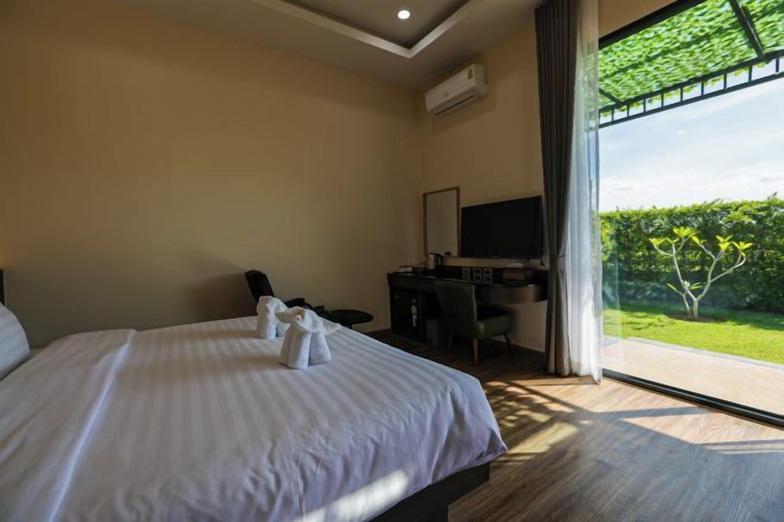 Habitación de hotel con una cama grande, un escritorio, una silla y un televisor. Se ve follaje verde a través de la ventana.