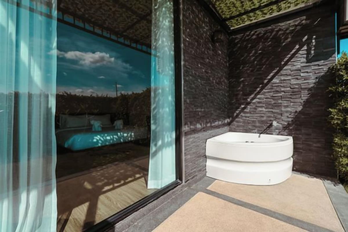 A modern bathroom with a large glass door, a white bathtub, and a stone wall.
