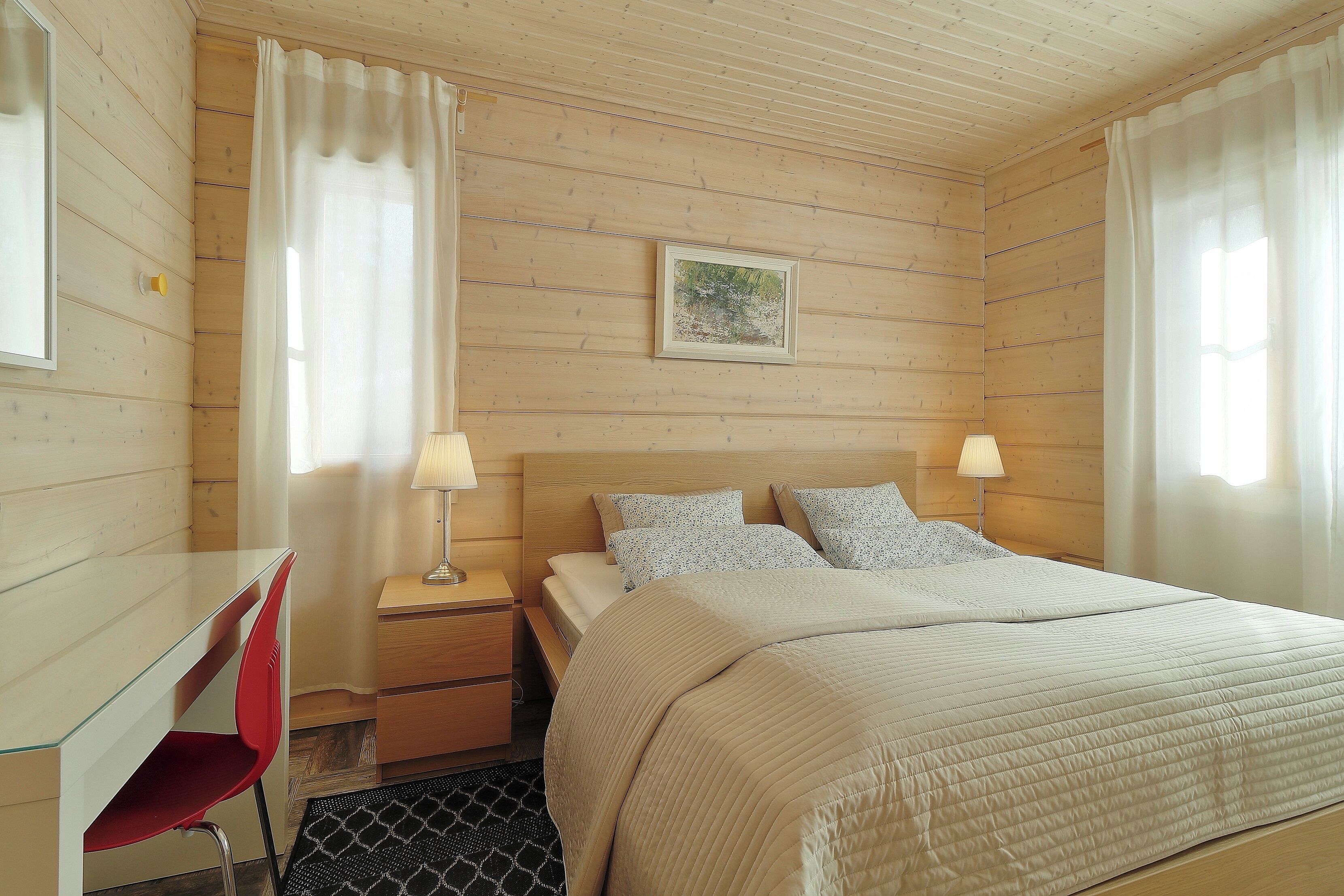 Un dormitorio con pared de madera, una cama con colcha color beige, dos lámparas de noche, una mesita con lámpara y una silla.