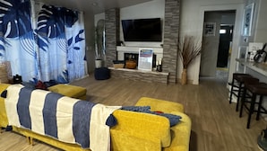 A modern living room with a yellow sofa, a ceiling fan, a flat-screen TV, and a fireplace. - Grand Sand Riverfront, private beach & boat dock (Parker)