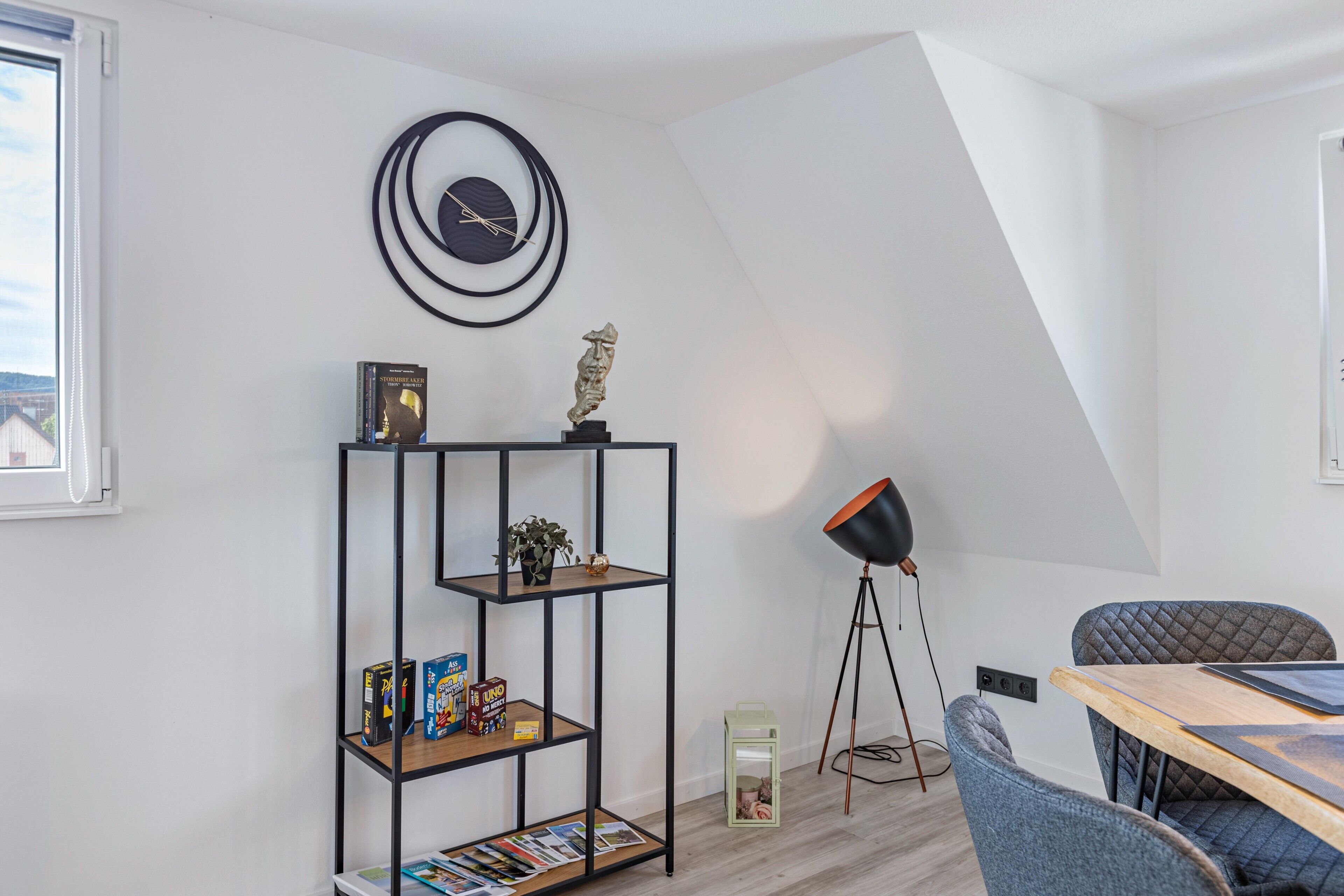 Una habitación moderna con una estantería metálica negra, un reloj de pared redondo, una lámpara de pie y una mesa de comedor con sillas.