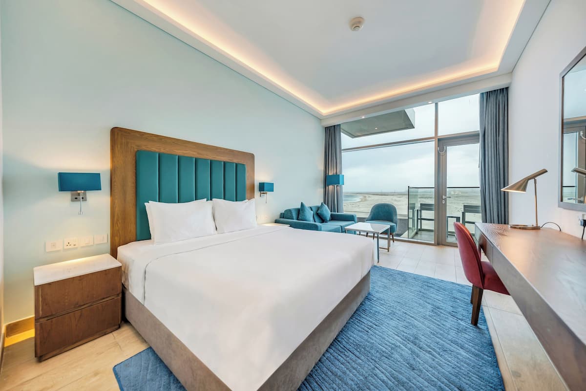A modern hotel room with a large bed, a desk, a chair, and a balcony with a view.