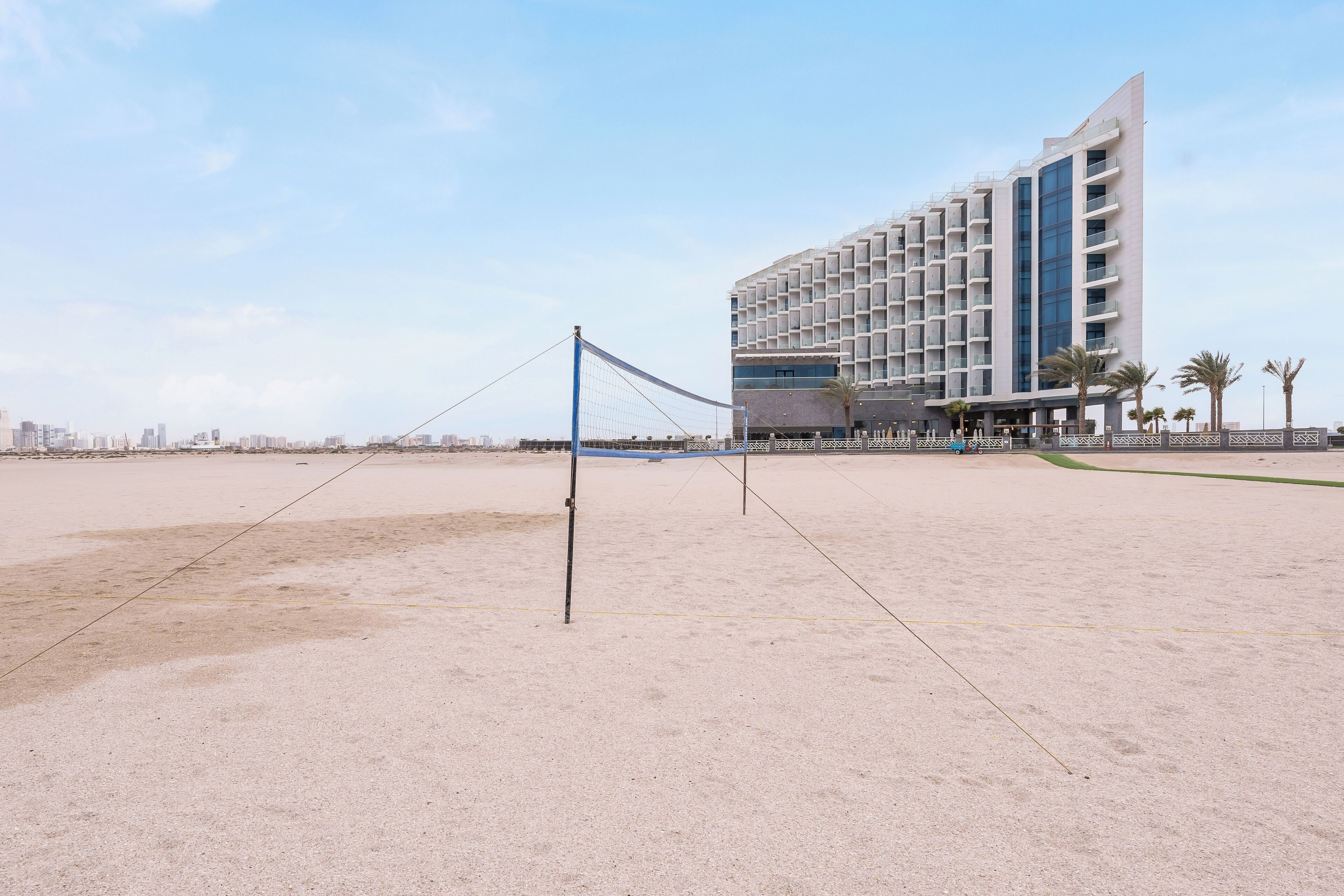 A beach volleyball court with a net, palm trees, and a modern hotel in the background.