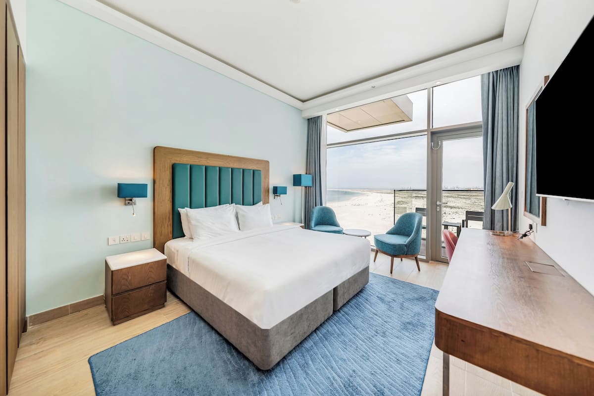 A hotel room with a large bed, a desk, a chair, and a view of the beach.