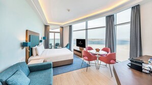 A hotel room with a bed, a sofa, a dining table with chairs, a TV, and large windows offering a view. - Sea Point Hotel (Dubai)