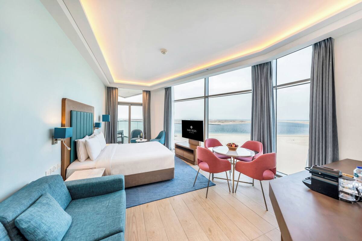 A hotel room with a bed, a sofa, a dining table with chairs, a TV, and large windows offering a view.