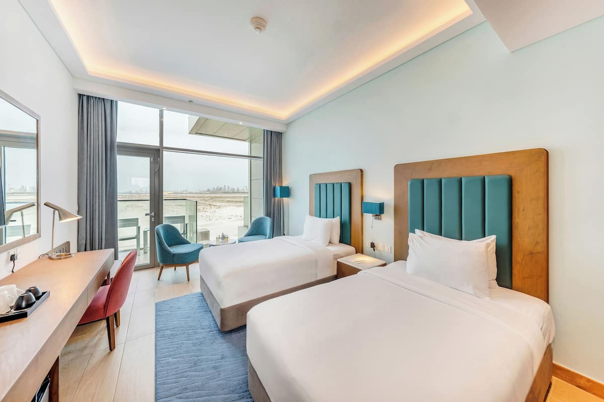 A hotel room with two beds, a desk, a chair, a lamp, and a view of the city.