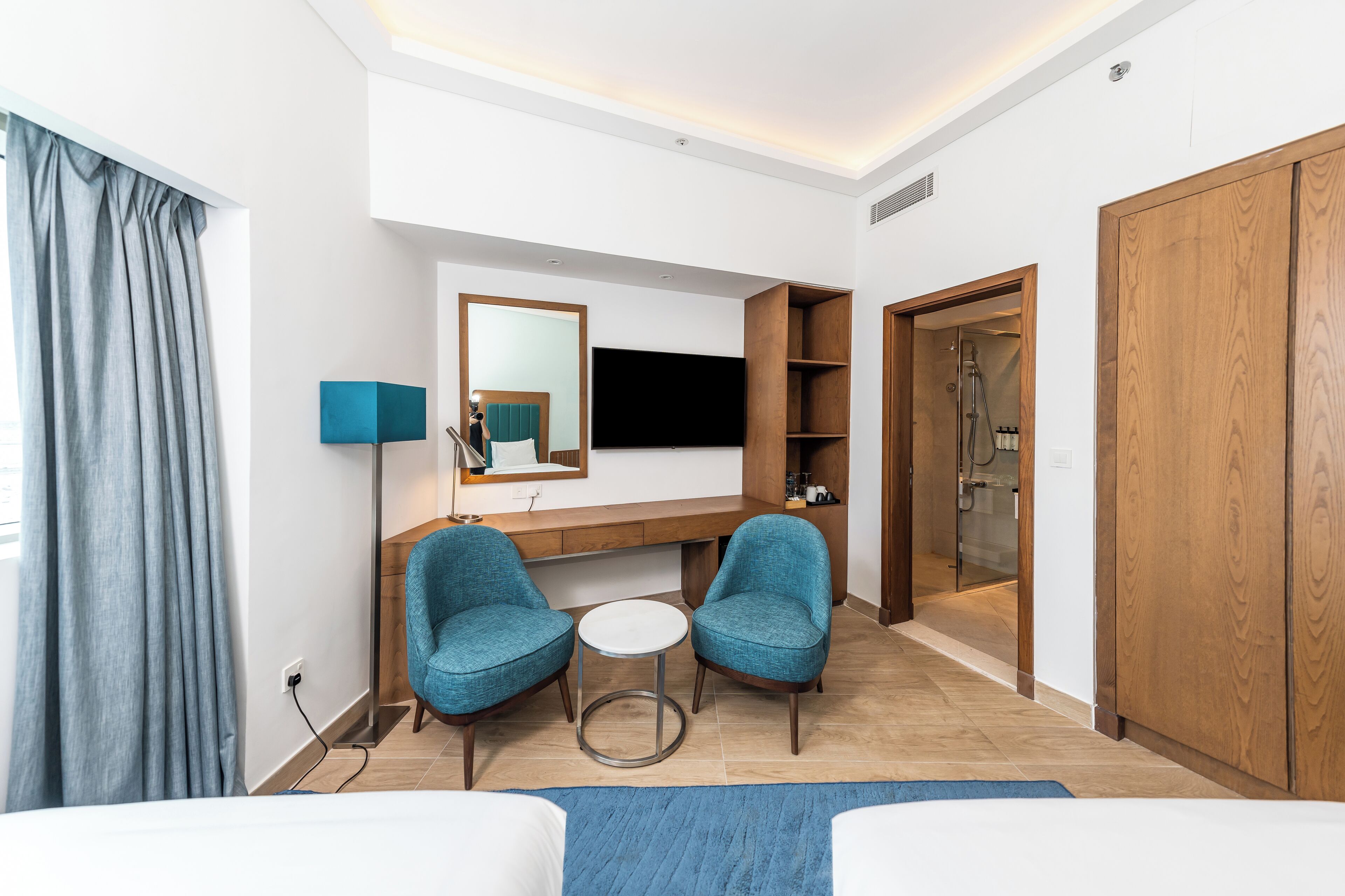 A hotel room with a bed, two armchairs, a small round table, a TV mounted on the wall, a wooden wardrobe, and a shower area.