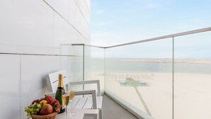 A balcony with a white table, a wicker fruit basket, a bottle of champagne, and a glass of wine. The view includes a sandy beach and a clear sky. - Sea Point Hotel (Dubai)