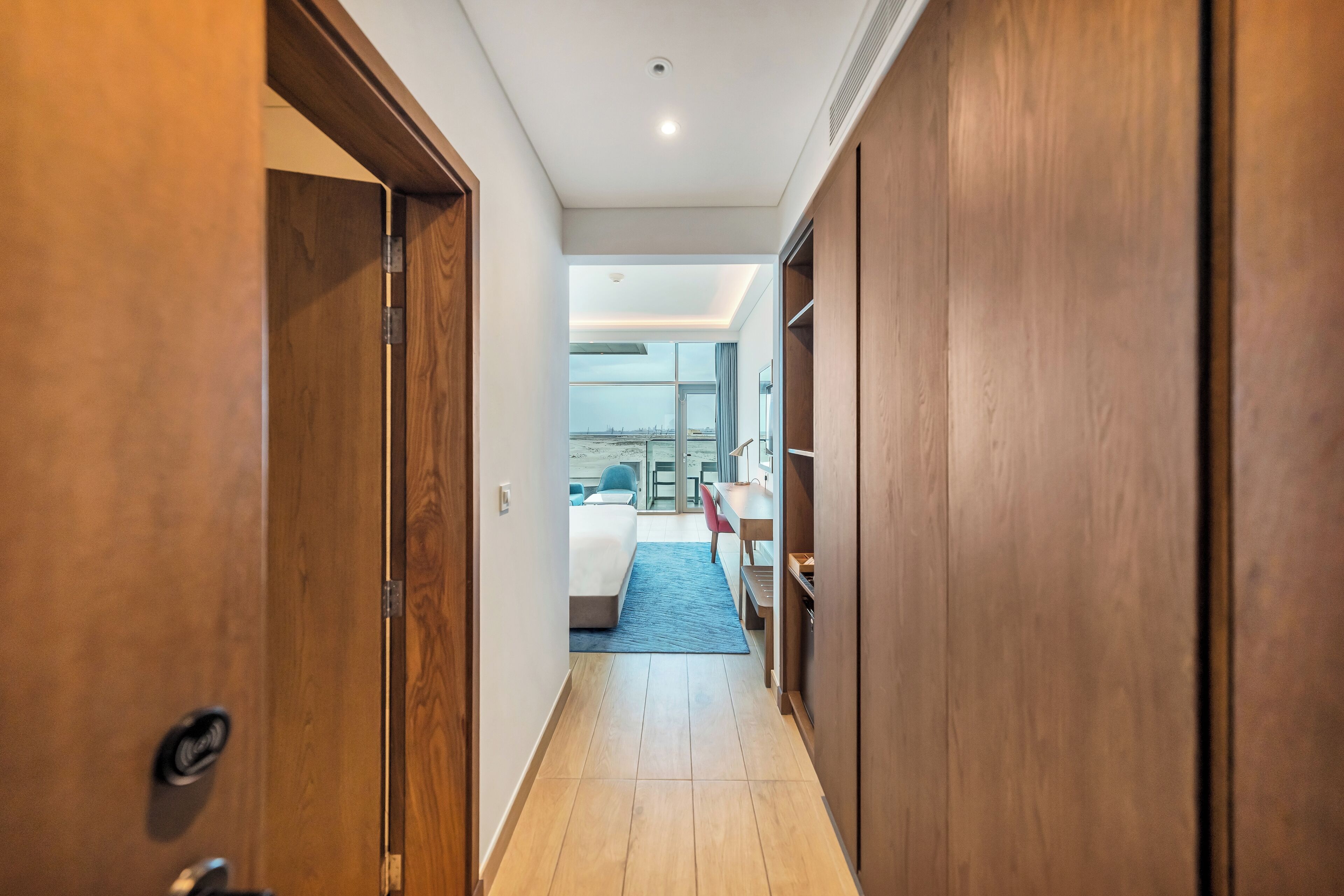 A hotel hallway with wooden doors, a bed with a blue bedspread, and a desk with a chair.