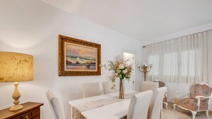 Een eetgedeelte met een tafel, stoelen, een lamp, een lijst met een schilderij en een vaas met bloemen.