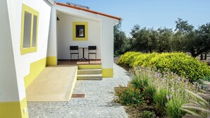A building with a yellow and white exterior, a small outdoor seating area with two tables and chairs, and a garden with yellow flowers and greenery. - CASA DAS COLMEIAS A haven of tranquillity, where nature reveals all its beauty. (Distrito de Évora)