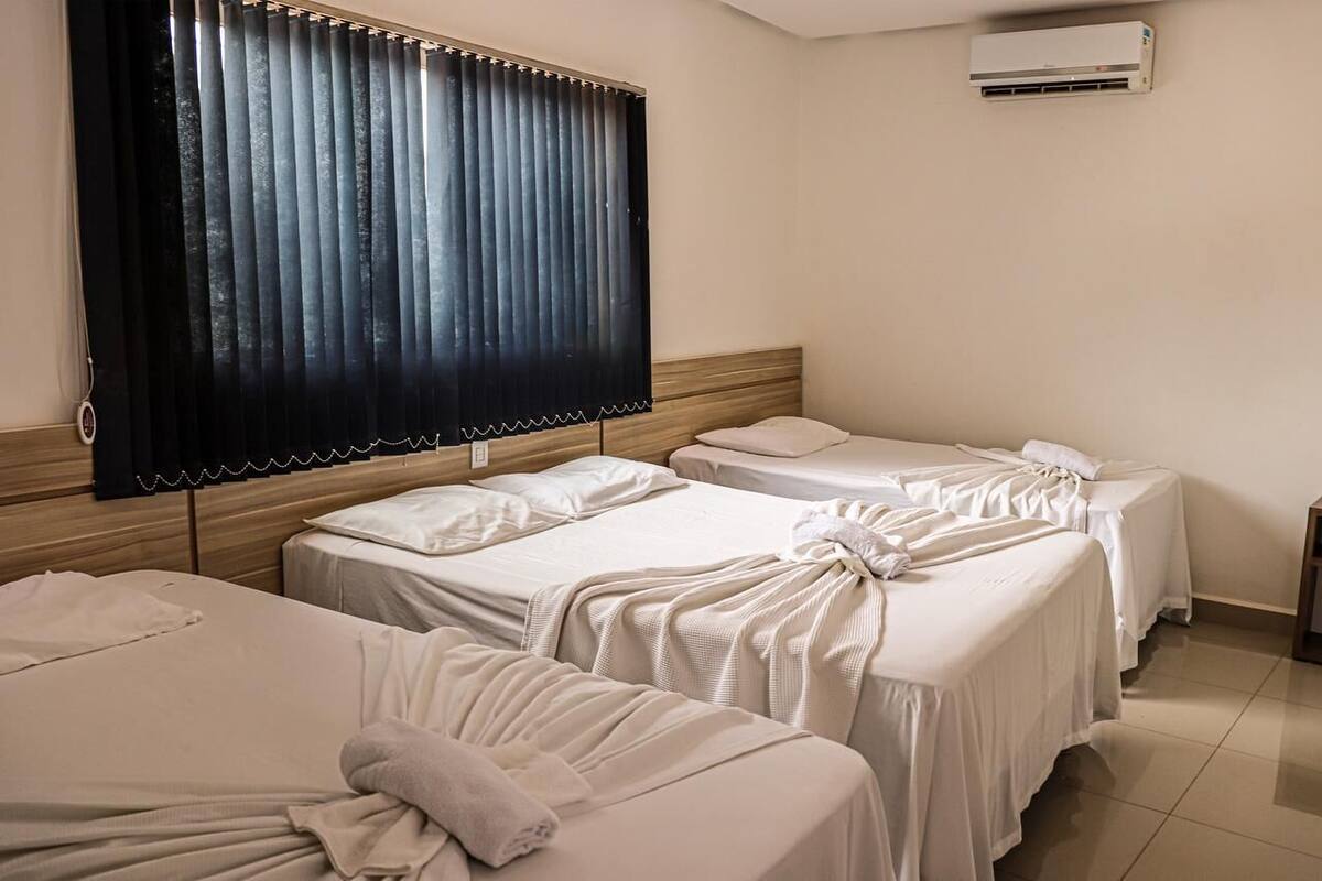 Ein Hotelzimmer mit drei Betten, einem hölzernen Kopfteil, einem großen Fenster mit vertikalen Jalousien und einer Klimaanlage an der Wand.