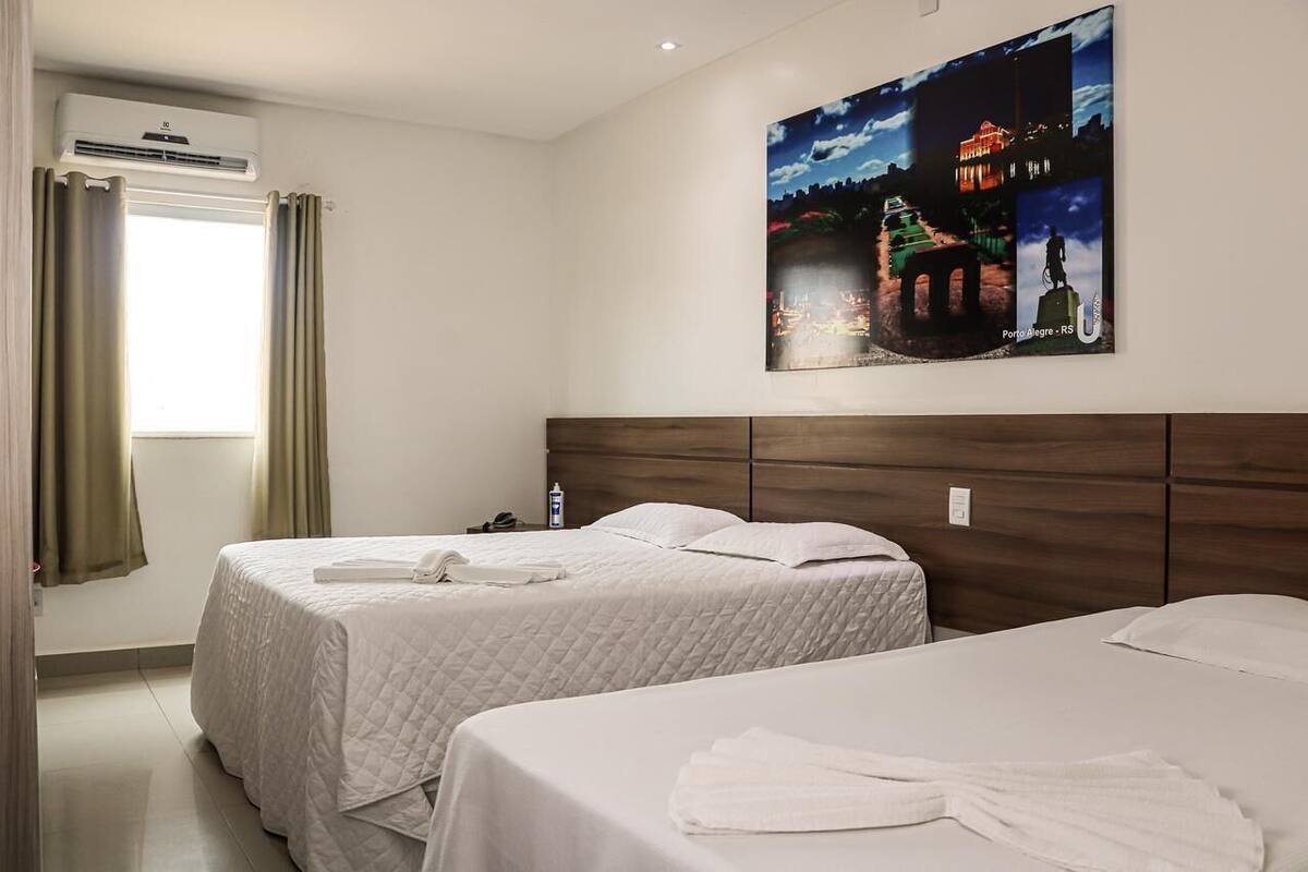 Ein Hotelzimmer mit zwei Betten, einem Stadtbild als Gemälde, einer Klimaanlage und einem Fenster mit Vorhängen.