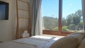 Une chambre avec une grande fenêtre, une échelle en bois, un téléviseur à écran plat et un lit avec une literie blanche.