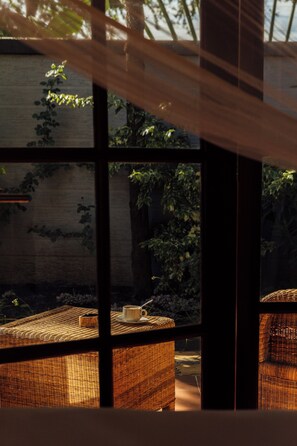 A wicker chair with a cup and saucer on a woven table, a window with a view of greenery, and a brick wall. - Shoki Shoki Lodge (Arusha)