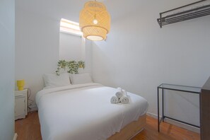 Una cama bien tendida con toallas enrolladas, una planta en la mesita de noche y una lámpara colgante.