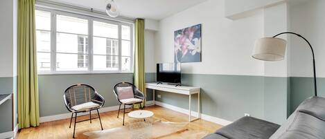 Ett modernt vardagsrum med en soffa, ett skrivbord, en stol, en tv och ett stort fönster med gardiner.