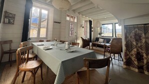 Un coin repas avec une table dressée pour quatre personnes, entourée de chaises en bois. On aperçoit un banc au fond de la pièce, et celle-ci est dotée de hauts plafonds avec des poutres apparentes.