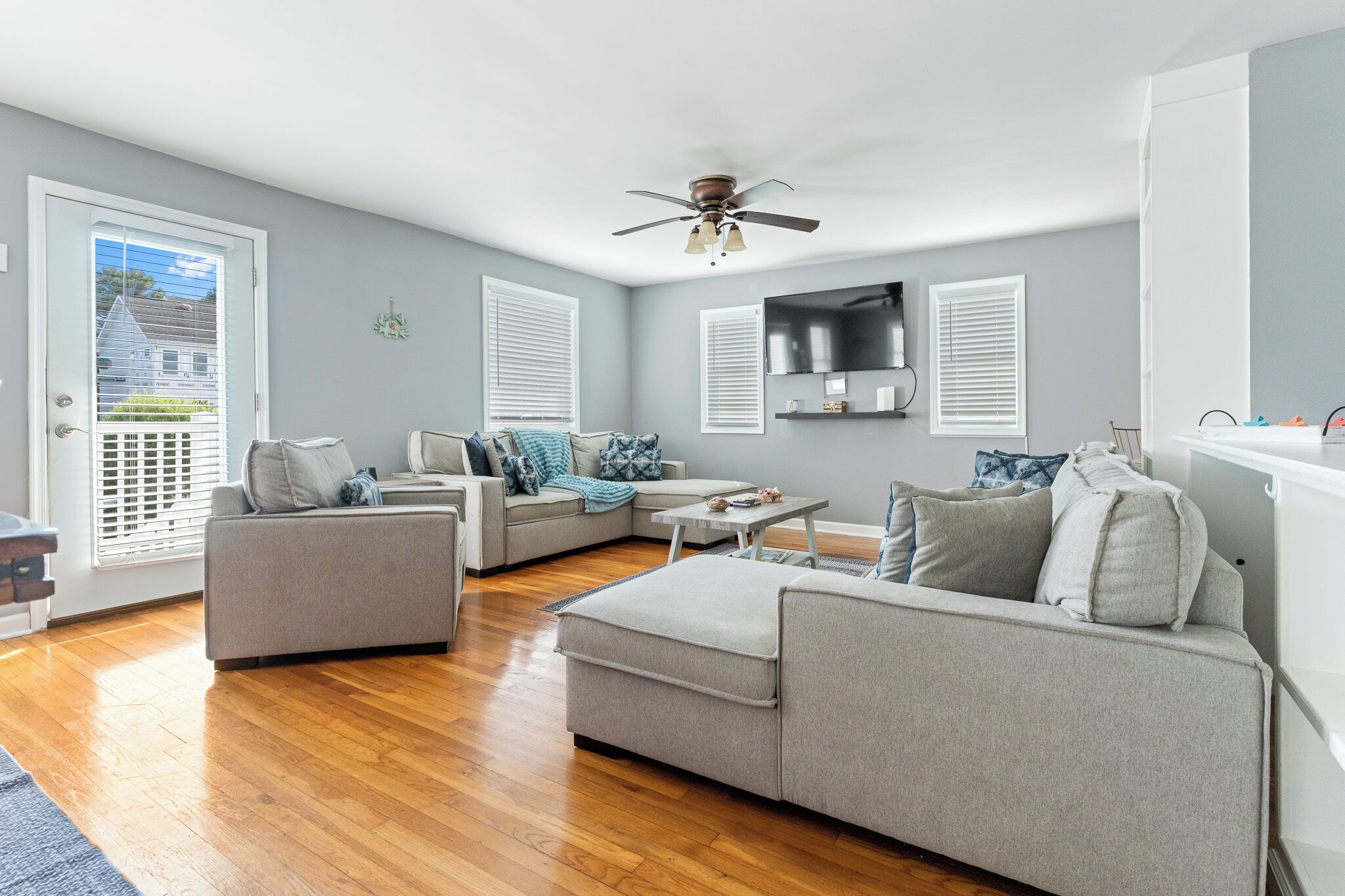 A modern living room with grey sofas, a ceiling fan, and a flat-screen TV mounted on the wall.