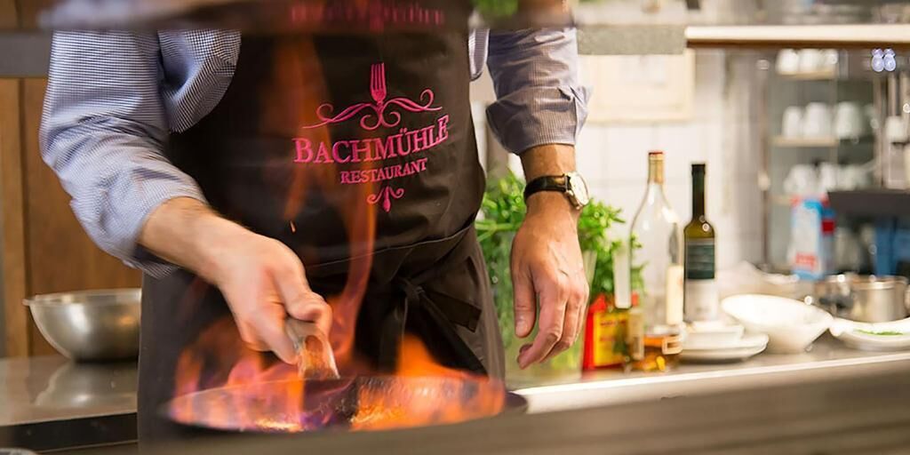 A person in a kitchen wearing a black apron with "BACHMÜHLE RESTAURANT" printed on it, cooking on a stove.