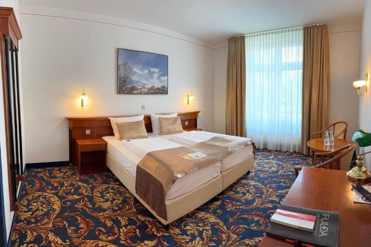 A hotel room with a large bed, two bedside tables, a desk with a chair, a window with curtains, and a painting on the wall.