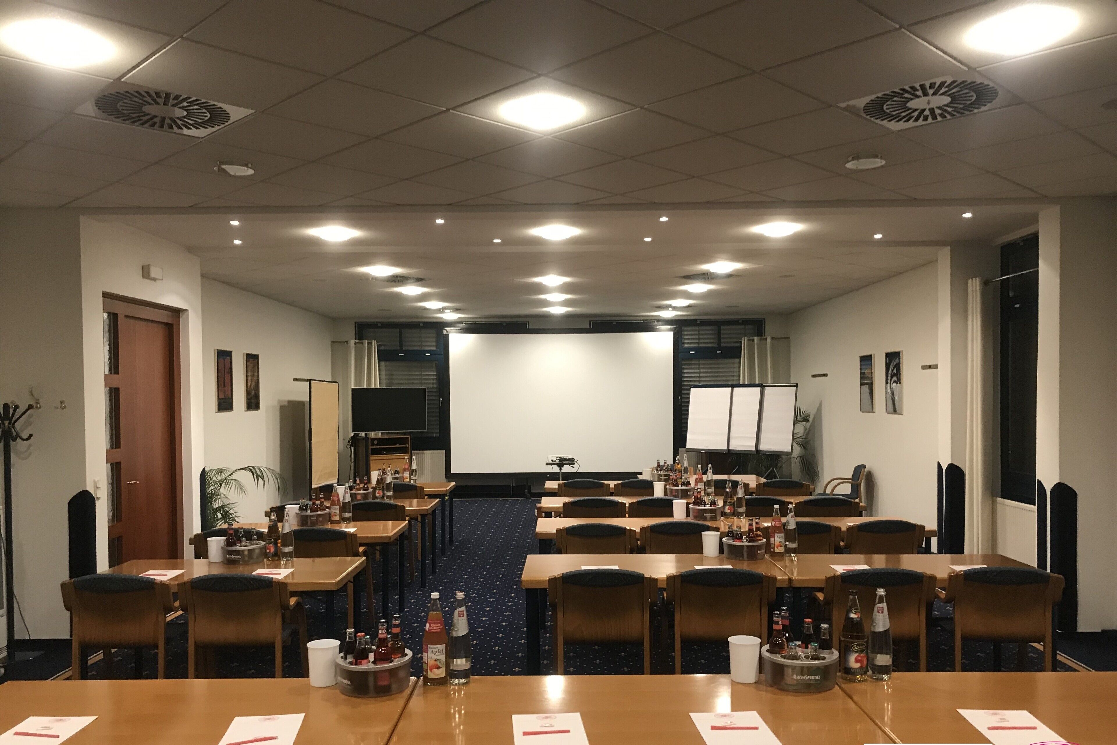 Un salón de conferencias con varias mesas, sillas y una gran pantalla al frente.
