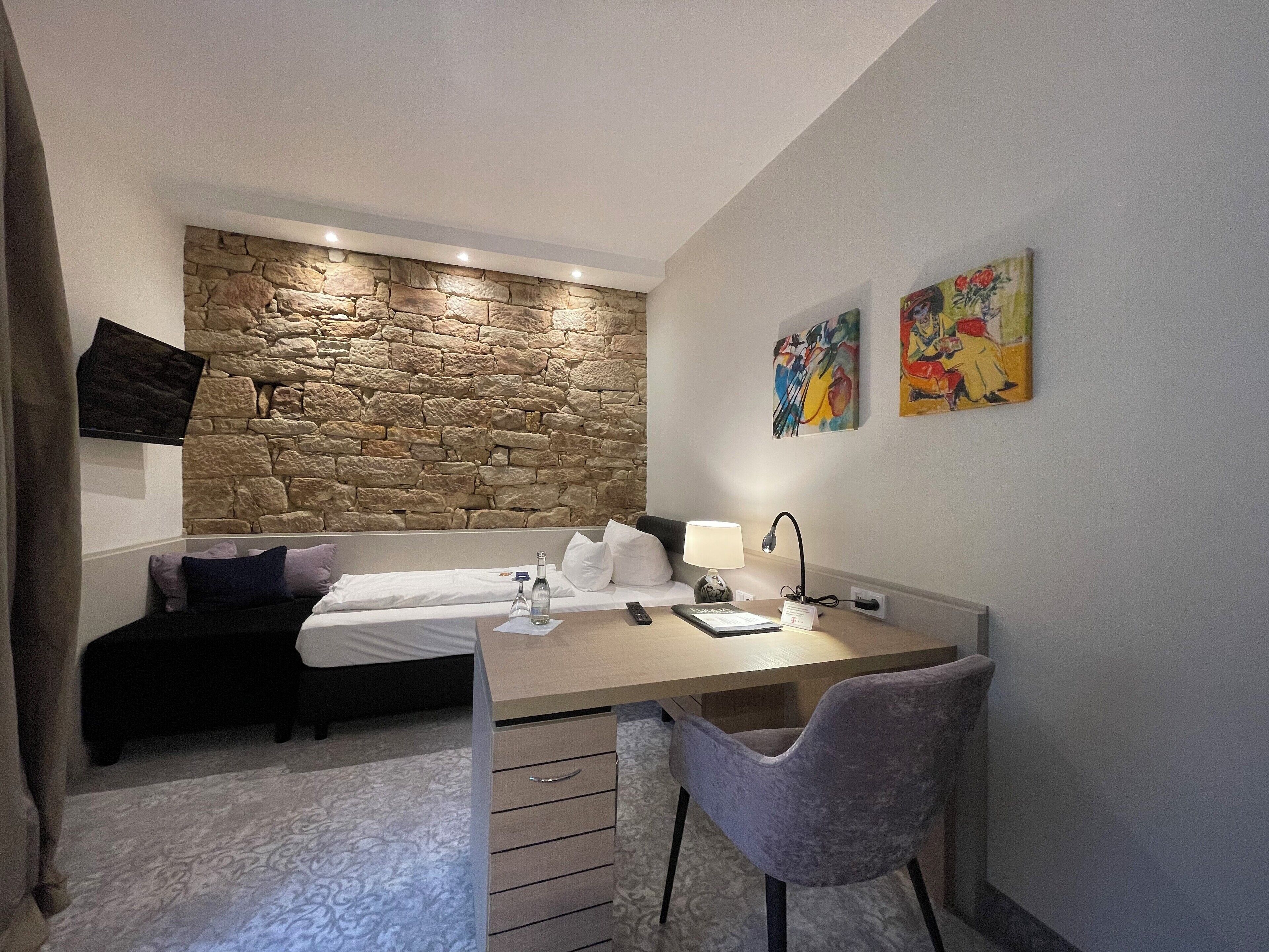Une chambre d’hôtel compacte avec un mur décoratif en pierre, un lit, un bureau avec une chaise et des œuvres d’art accrochées au mur.