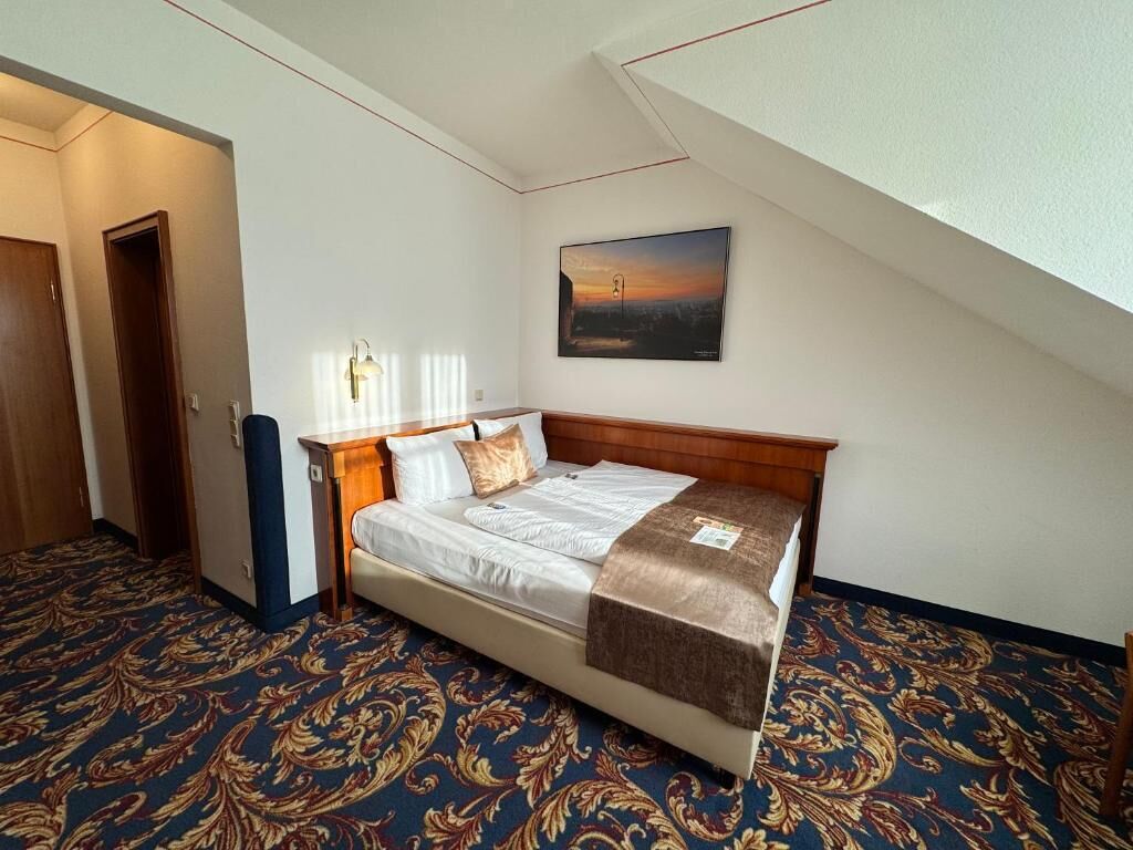 A hotel room with a bed, a nightstand, a lamp, a painting on the wall, and a patterned carpet.