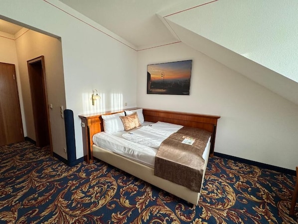 A hotel room with a bed, a nightstand, a lamp, a painting on the wall, and a patterned carpet.