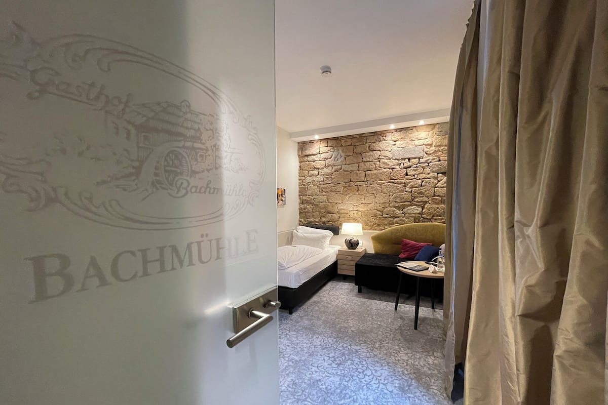 A hotel room with a stone wall, a bed with white linens, a small table with a lamp, and a chair.