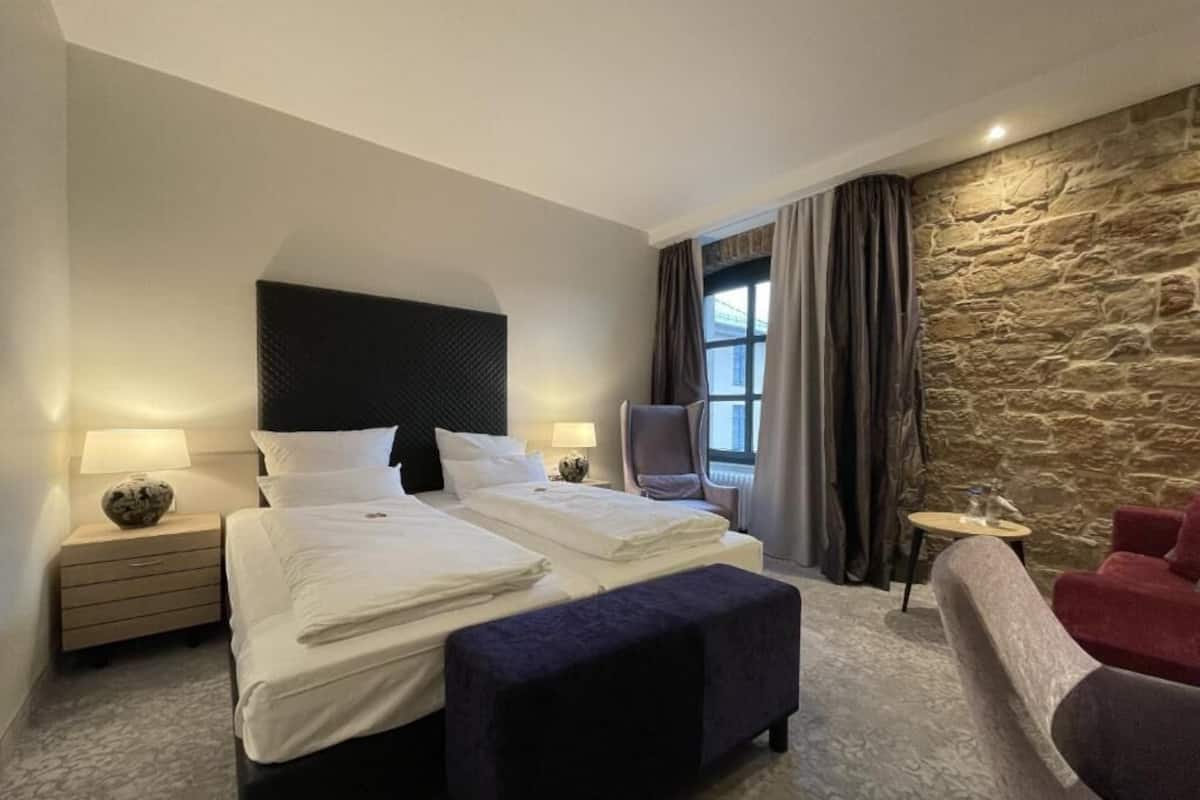 A hotel room with a large bed, a chair, a small table, and a stone wall.