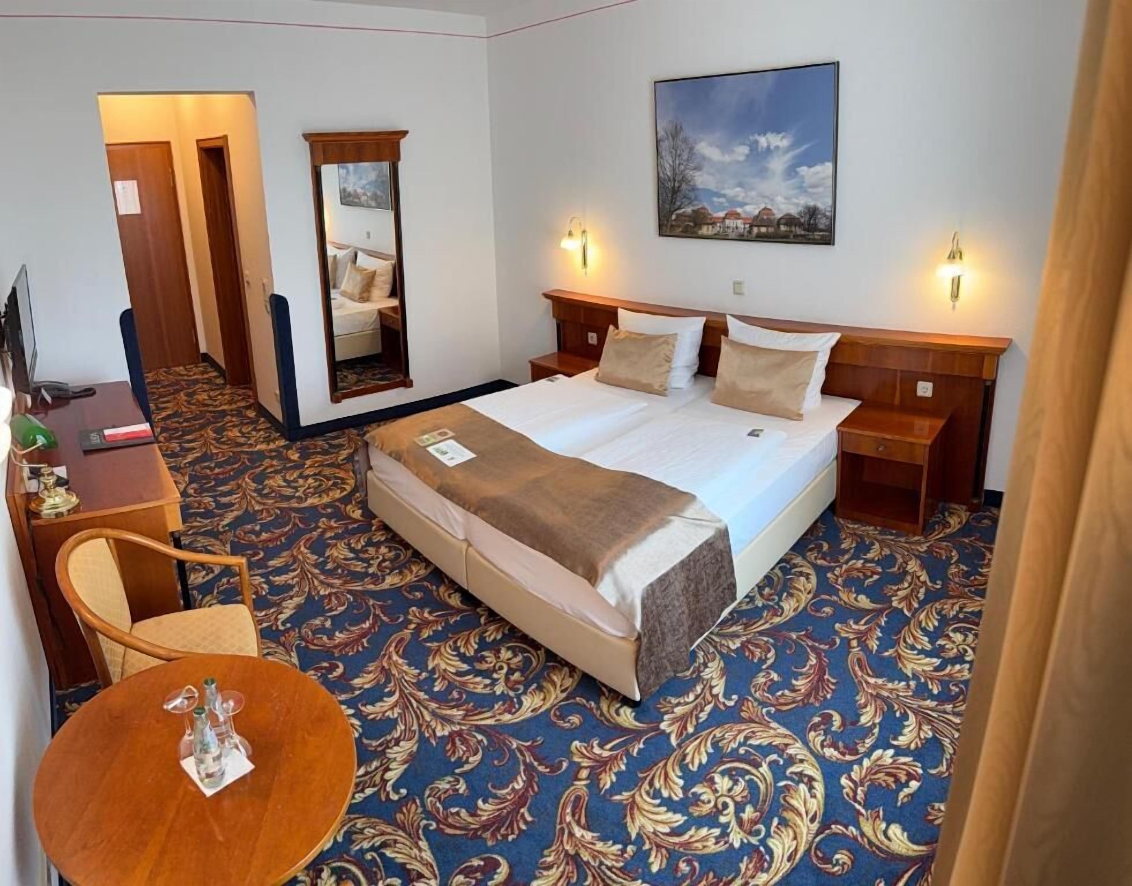 A hotel room with a large bed, two bedside tables, a desk with a chair, a mirror, and a painting on the wall.