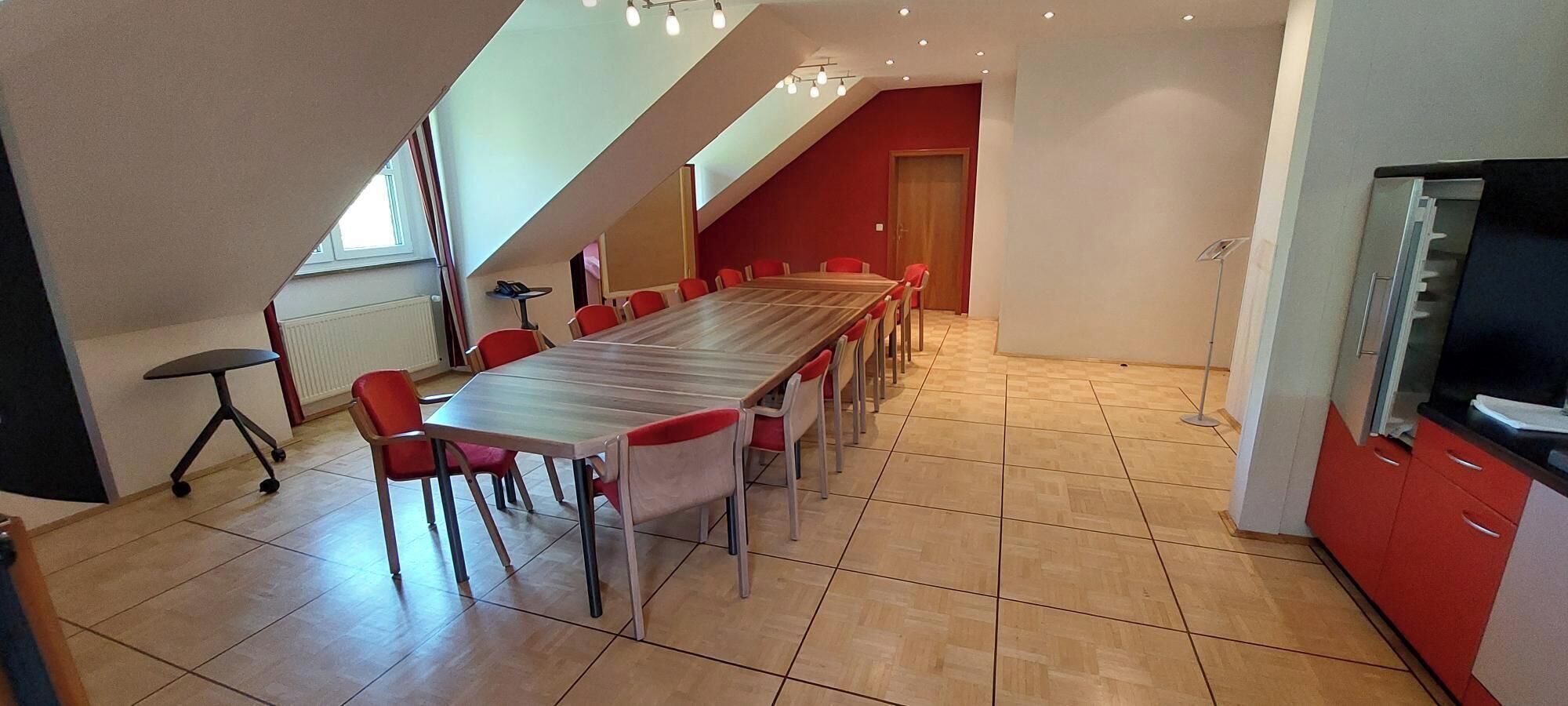 A dining area with a long table and red chairs, a kitchen with red cabinets, and a sloped ceiling with skylights.