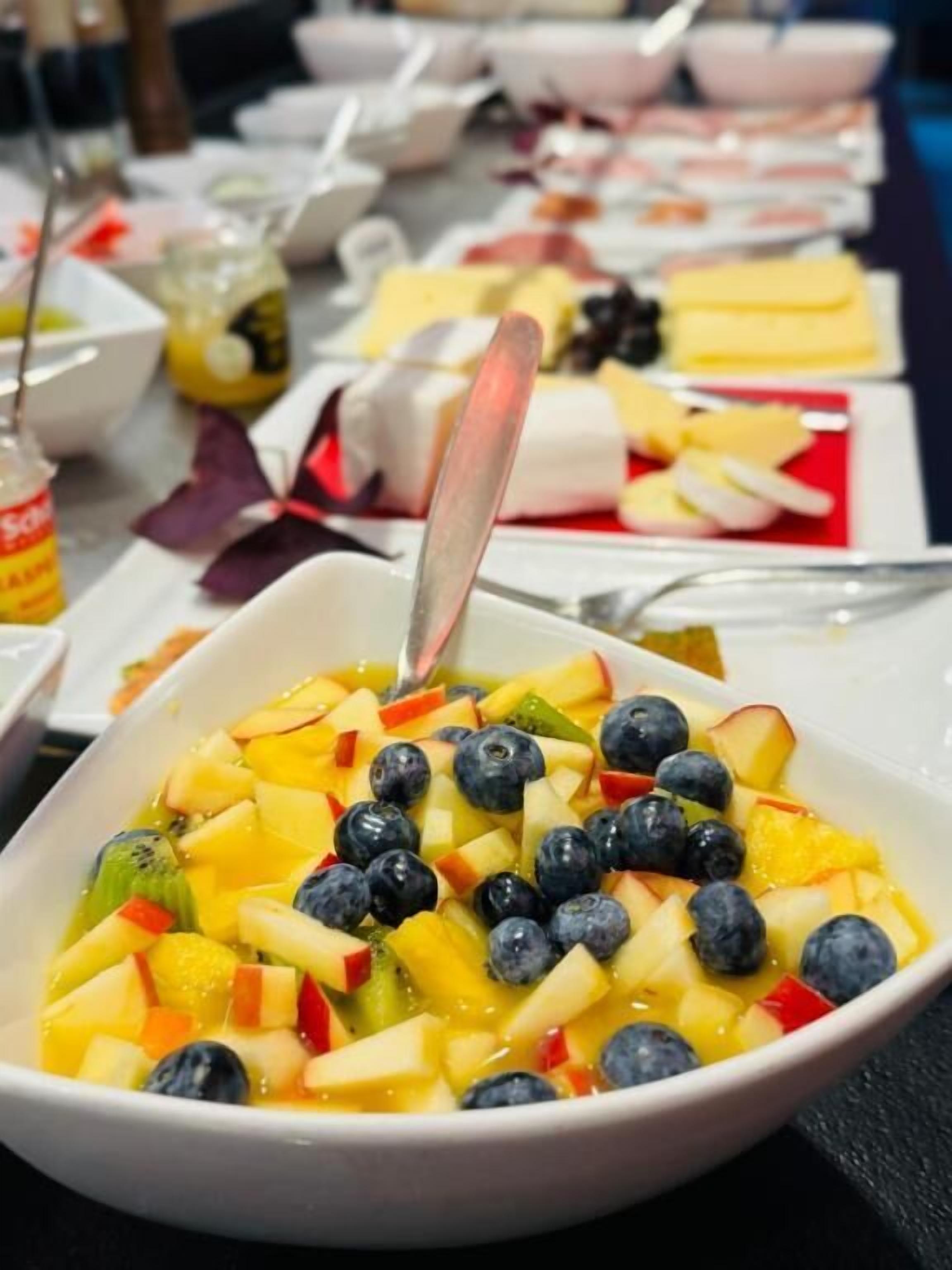 Una mesa de buffet con diversos platos, incluyendo un tazón de ensalada de frutas con arándanos, manzanas y kiwi.