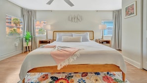 A bedroom with a large bed, a ceiling fan, two bedside tables with lamps, a floral-patterned rug, and a view of buildings through the window. - 2BR Waterview Suite on Clearwater Beach w/ Pool (Clearwater)