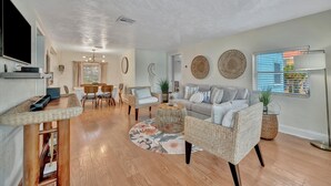 A spacious living room with a large TV, wicker furniture, a round coffee table, and a dining area with chairs and a table. - 2BR Waterview Suite on Clearwater Beach w/ Pool (Clearwater)