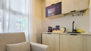 Una kitchenette compacta con microondas, cafetera y hervidor. Un sofá se encuentra cerca de una ventana con cortinas traslúcidas.