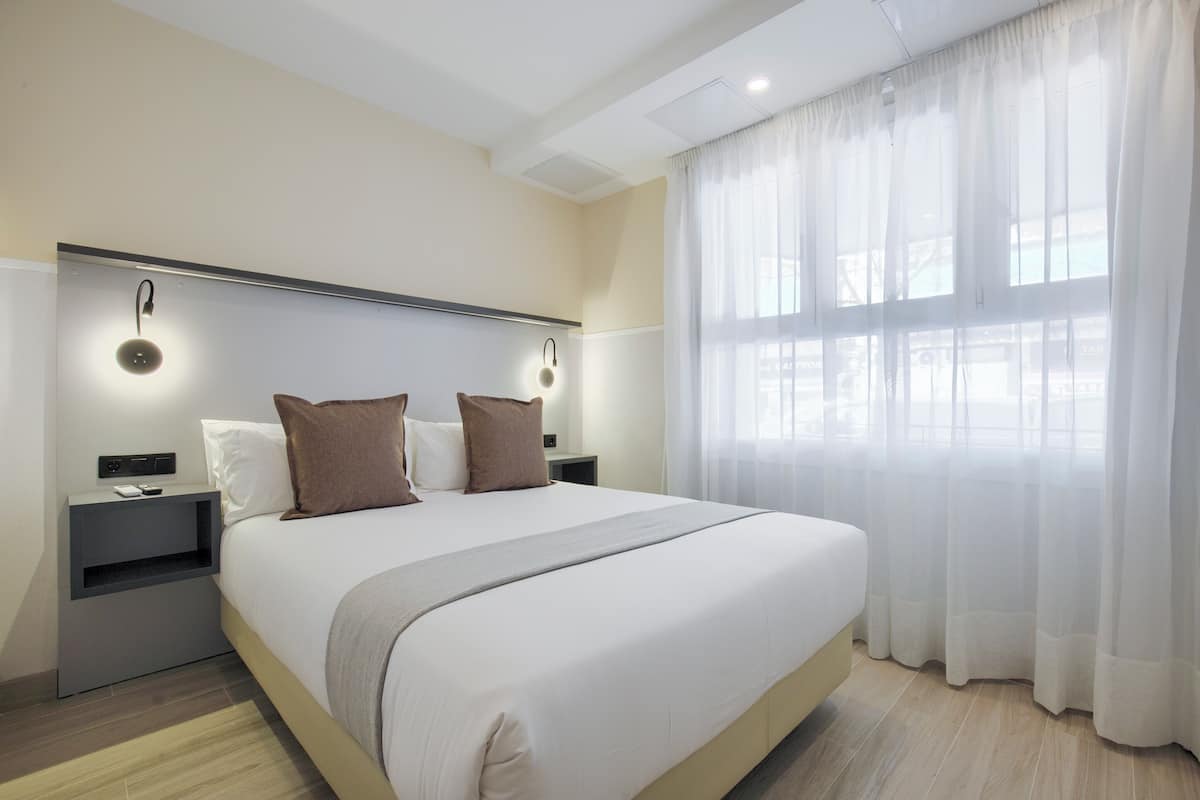 Una habitación de hotel moderna con una cama grande, mesitas de noche y una ventana con cortinas translúcidas.
