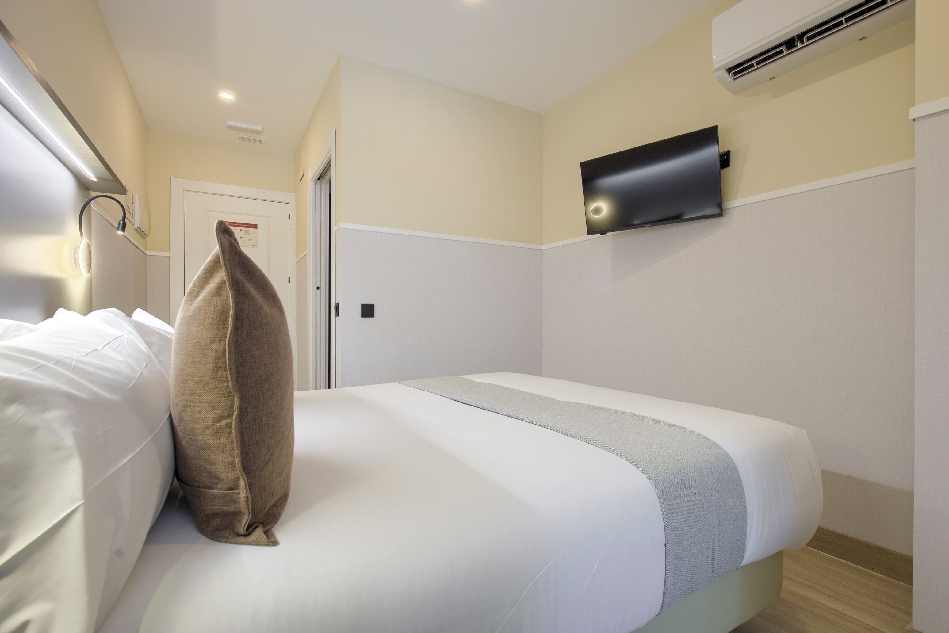 Una cama bien hecha con ropa de cama blanca y gris, un almohadón marrón y un televisor de pantalla plana montado en la pared.