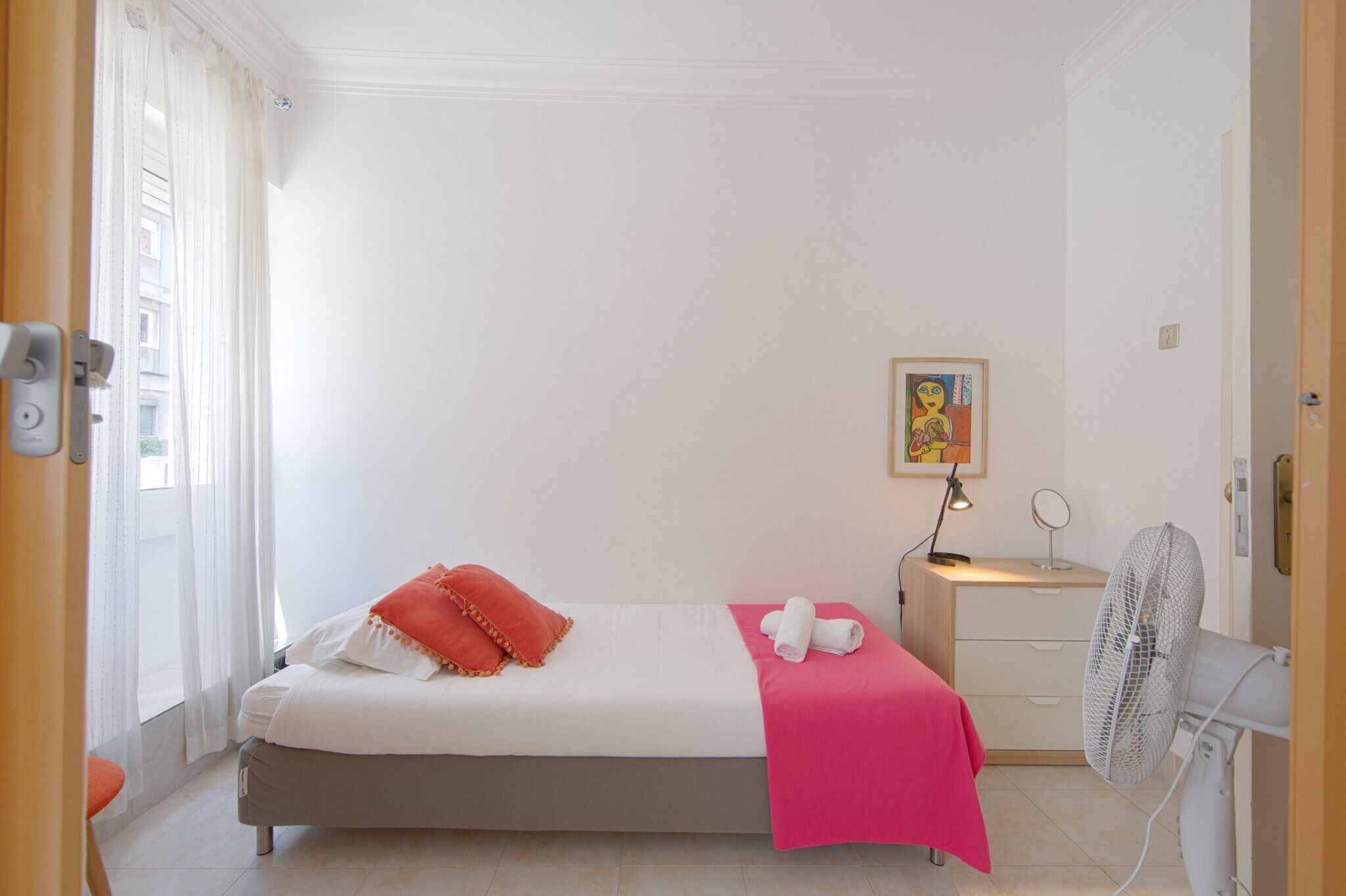 Une petite chambre à coucher, bien rangée, avec un lit, une table de chevet, un ventilateur et une fenêtre avec des rideaux.