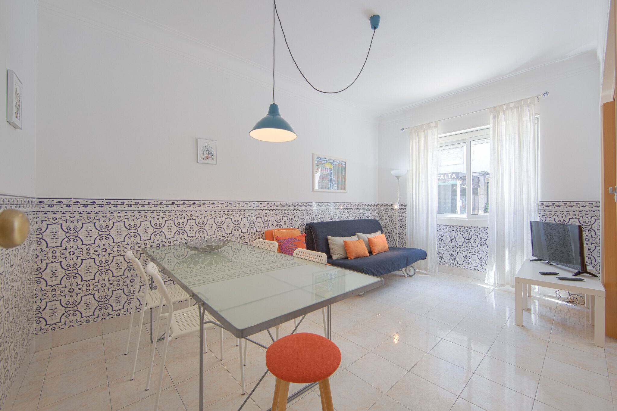 Un salon moderne avec une table à manger en verre, un canapé agrémenté de coussins colorés, un téléviseur posé sur un meuble et des murs à motifs.