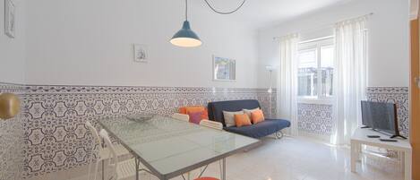 Un salon moderne avec une table à manger en verre, un canapé agrémenté de coussins colorés, un téléviseur posé sur un meuble et des murs à motifs.