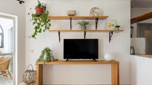 A living room with a TV on a wooden stand, shelves with plants and decorative items, and a wicker chair by the window. - Cascais Ocean View by Homing (Cascais)