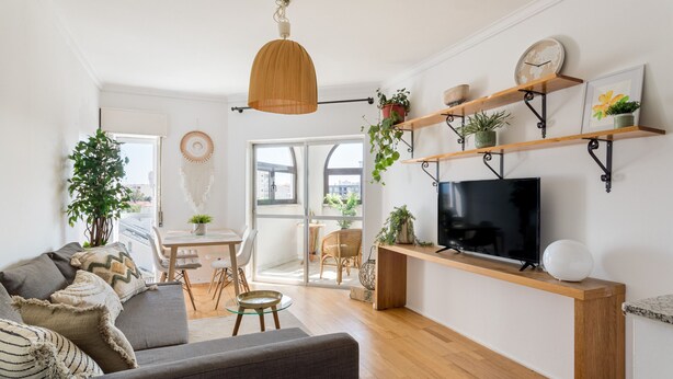 A modern living room with a sofa, TV, wooden shelves with plants, and a dining area with chairs and a table. - Cascais Ocean View by Homing (Cascais)