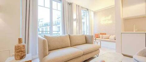 Un salon moderne avec un canapé beige, une table basse ronde et une vue sur le paysage urbain, visible à travers de grandes fenêtres.