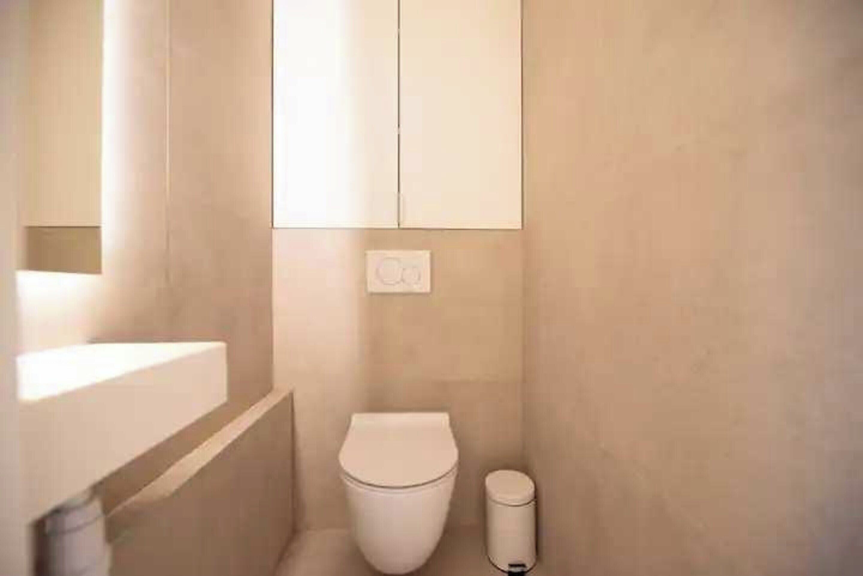 A small, compact bathroom with a toilet, a trash bin, and a wall-mounted light fixture.