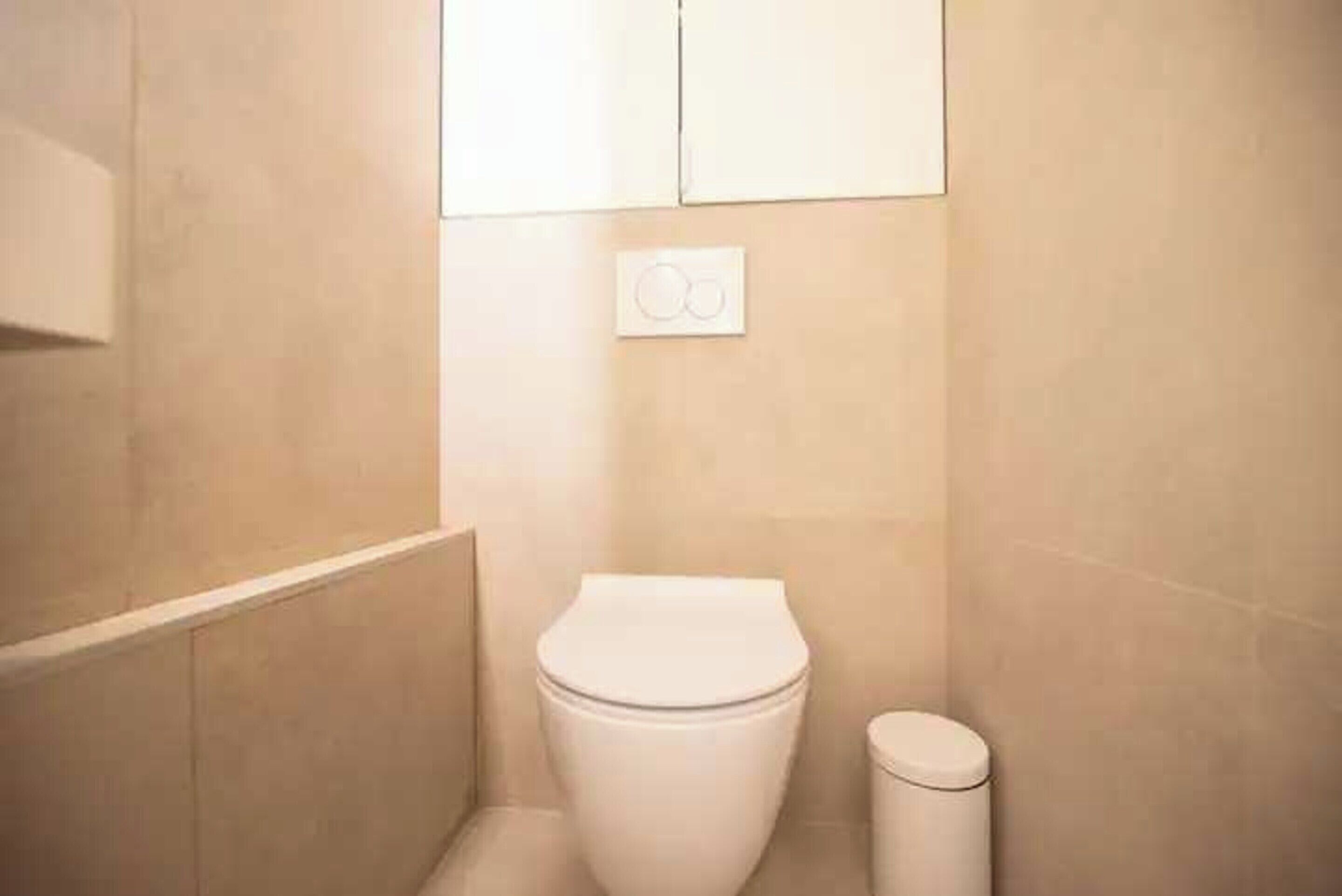 A small, clean bathroom with a toilet, a trash bin, and a wall-mounted light switch.