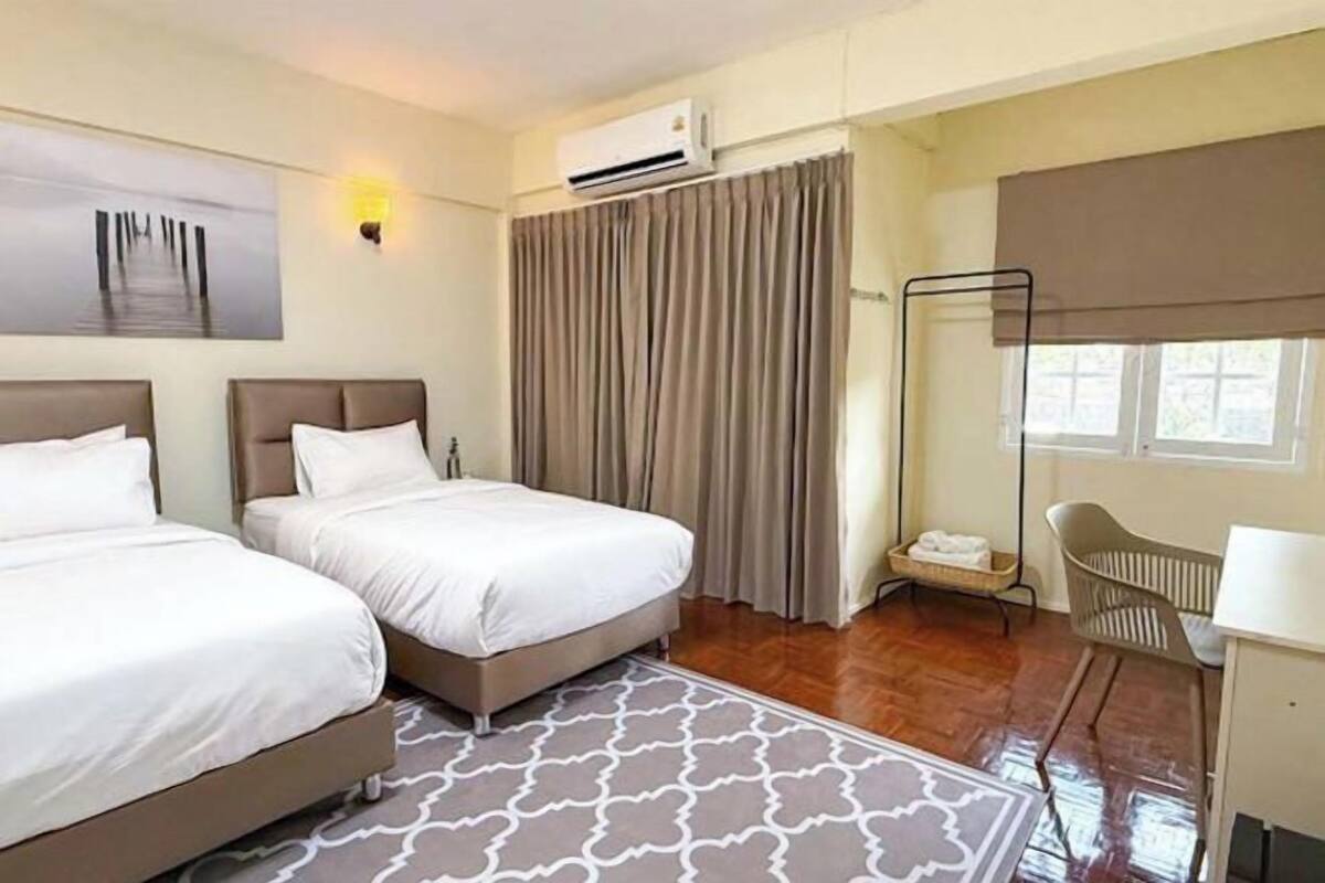 A hotel room with two beds, a desk, a chair, an air conditioning unit, a window with curtains, and a wall-mounted artwork.