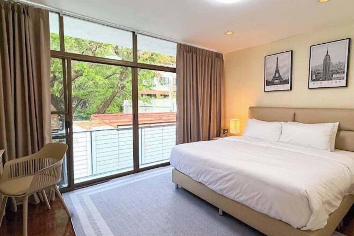 A hotel room with a large bed, a chair, a window with curtains, and two framed pictures on the wall.